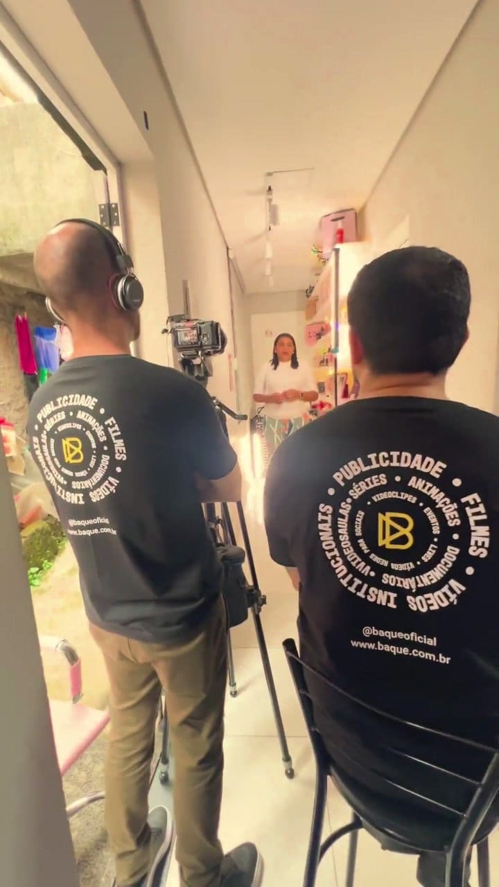 Dois integrantes da equipe da Baque de costas, com camisetas da produtora, enquanto a entrevistada fala de frente para a câmera