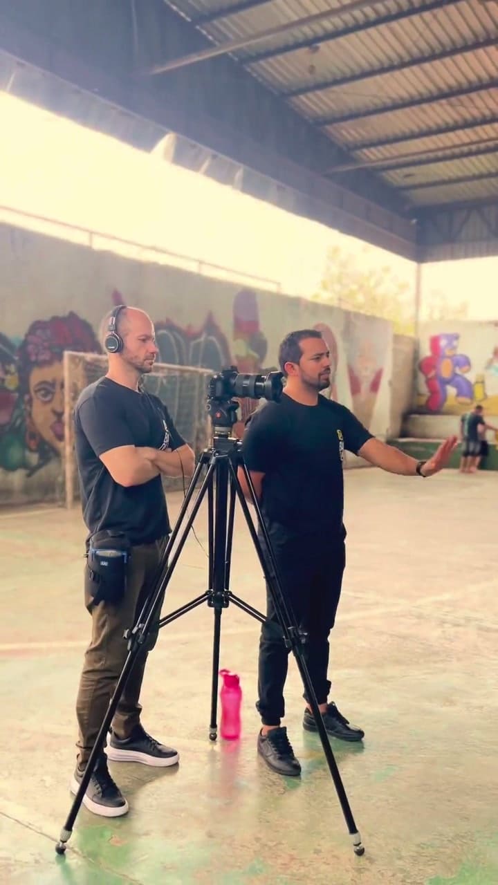 Dois integrantes da equipe da Baque atrás da câmera montada em tripé, numa quadra coberta com muros grafitados