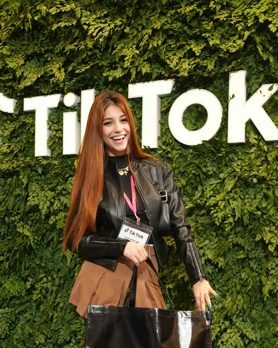 Convidada com credencial do TikTok posa diante do painel de folhagem com o logo da marca, produção Baque